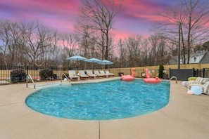 A swimming pool with lounge chairs and umbrellas, surrounded by a fence and trees. - Greensboro Getaway Villa (Greensboro)