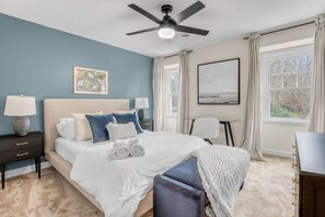 A bedroom with a bed, two bedside tables, a ceiling fan, a window with curtains, and artwork on the walls. - Greensboro Getaway Villa (Greensboro)
