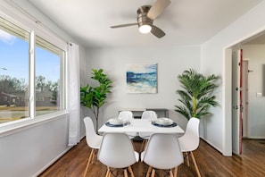 Un coin repas avec une table dressée pour quatre personnes, des chaises blanches, un ventilateur de plafond et un tableau accroché au mur.