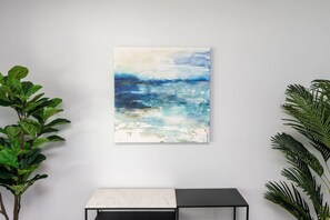 Una habitación con pared blanca, un cuadro de una escena oceánica azul, un banco blanco y negro, y plantas verdes a ambos lados.