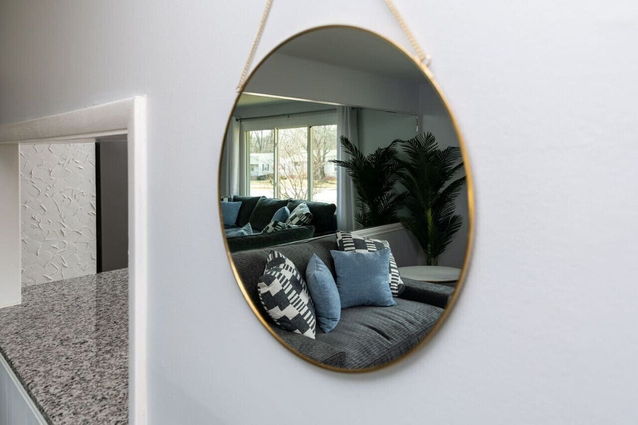A modern living room with a large oval mirror on the wall, a grey sofa with patterned cushions, and a large potted plant.