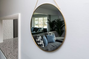 Una sala de estar moderna con un espejo ovalado grande en la pared, un sofá gris con cojines estampados y una planta grande en maceta.