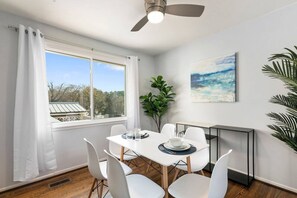 Comedor con una mesa dispuesta para cuatro, sillas blancas, ventilador de techo, ventana grande con cortinas, un cuadro en la pared y una planta.