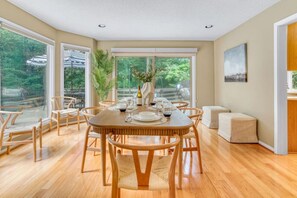 A dining area with a round wooden table set for six, surrounded by six chairs. Large windows provide a view of greenery outside. - Arlington Oasis 7 Br (Jamestown)