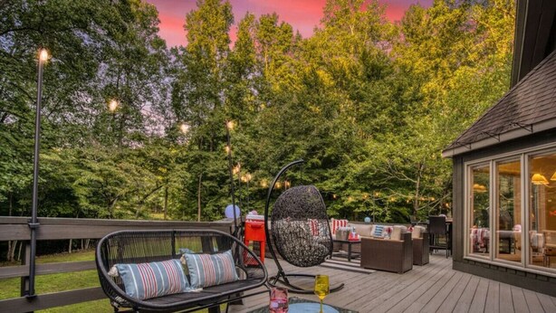 An outdoor deck with seating, a glass-enclosed structure, and trees in the background. - Arlington Oasis 7 Br (Jamestown)