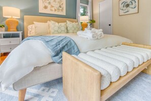 A neatly made bed with a blue blanket and white towels, a bedside table with a lamp, and a wall with floral artwork. - Arlington Oasis 7 Br (Jamestown)