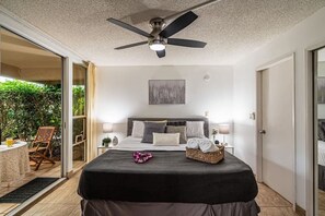 Ein Schlafzimmer mit Bett, Deckenventilator, Nachttisch mit Lampe, Fenster und Blick ins Freie.