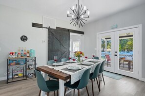 A dining area with a wooden table set for four, teal chairs, a chandelier, and a view of the outdoors through a glass door. - Renovated 6BR Near Grand Lake Fire Pit & Games (Grove)