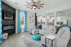 A modern living room with a fireplace, grey sofa, teal ottoman, and a kitchen in the background. - Renovated 6BR Near Grand Lake Fire Pit & Games (Grove)