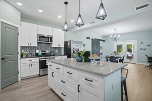 A modern kitchen with white cabinets, a granite countertop, and a central island. There is a dining area with a table and chairs visible through an open door. - Renovated 6BR Near Grand Lake Fire Pit & Games (Grove)