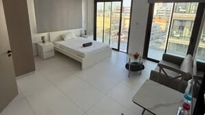 Une chambre moderne avec un grand lit, une table de chevet, une petite table avec une chaise, et une vue sur la ville grâce à de grandes fenêtres.