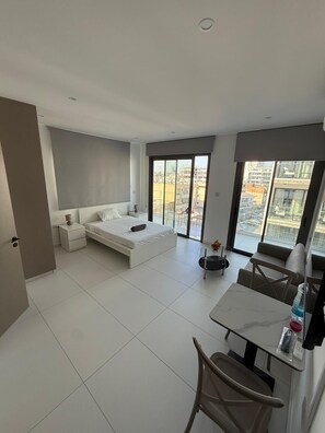 Ein modernes Schlafzimmer mit einem großen Bett, einem Nachttisch, einem kleinen Tisch mit Stuhl und Blick auf die Stadt durch große Fenster.