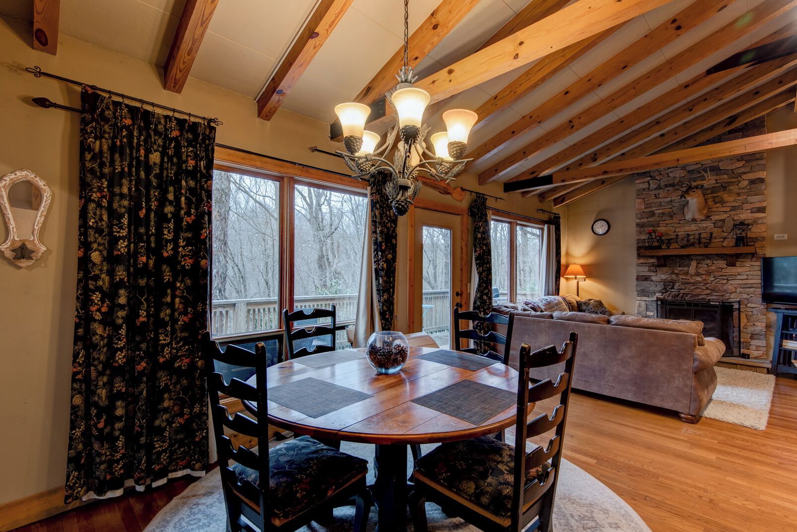A cozy living room with a stone fireplace, wooden beams, and a large window overlooking a deck.