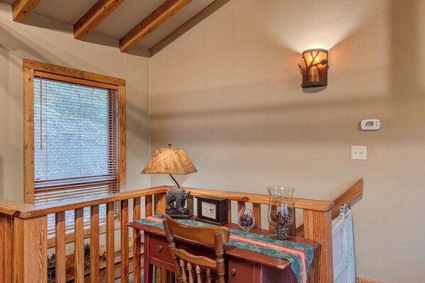 A wooden staircase with a railing, a table with chairs, a lamp, a wall-mounted light fixture, and a window with blinds. - Appalachian Retreat (Blowing Rock)