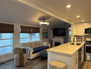 A modern kitchen with a built-in island, a flat-screen TV above the fireplace, and a dining area with a sofa and ottoman. - Blue Heron 447 (Jensen Beach)