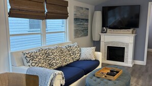 Ein Wohnzimmer mit Sofa, Hocker, Kamin, Fernseher und Deckenleuchte.