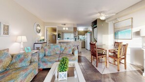 A living room with a sofa, a coffee table with a plant, a dining area with wooden chairs, and a kitchen in the background. - Summit 1013 (Panama City Beach)