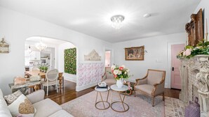 A spacious living room with a sofa, chairs, a coffee table, and a fireplace. There is a dining area with a table and chairs visible through an archway. - Pink House (Clarksville)
