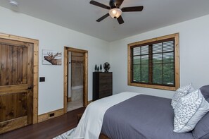 Une chambre à coucher comprenant un lit, un ventilateur de plafond, une fenêtre avec des stores, une porte en bois et un ventilateur fixé au mur.