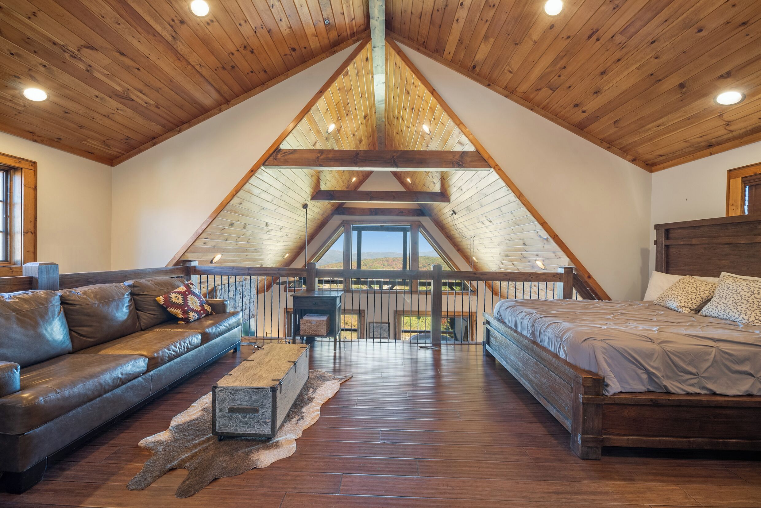 Ein geräumiges Schlafzimmer im Loft-Stil mit schrägem Dach, einem hölzernen Bett, einem Ledersofa und Blick ins Freie.