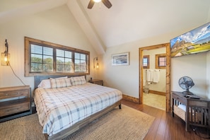 A bedroom with a large bed, wooden headboard, a window with blinds, a bedside table, a nightstand, a wall-mounted lamp, a framed picture, a mirror, a fan, and a television. - Peregrine's Perch at Eagles Nest (Elk Park)