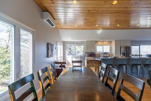 A spacious dining area with wooden flooring and a long table, leading to a kitchen with modern appliances and a view of the outdoors. - Alpenglow (Blowing Rock)