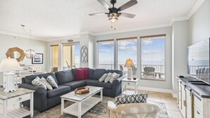 A living room with a gray sectional sofa, a white coffee table, and a ceiling fan. There are large windows with blinds, a dining area with a white table and chairs, and a kitchen area with a black countertop. - Gulf Crest 2005 (Panama City Beach)