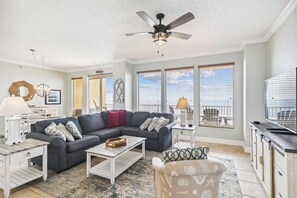 Ein Wohnzimmer mit grauem Ecksofa, einem weißen Couchtisch und einem Deckenventilator. Große Fenster mit Jalousien, ein Essbereich mit weißem Tisch und Stühlen sowie eine Küchenzeile mit schwarzer Arbeitsplatte.