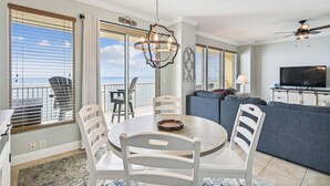 A dining area with a round table and white chairs, a balcony with a view, a living room with a sofa and TV, and a ceiling fan. - Gulf Crest 2005 (Panama City Beach)