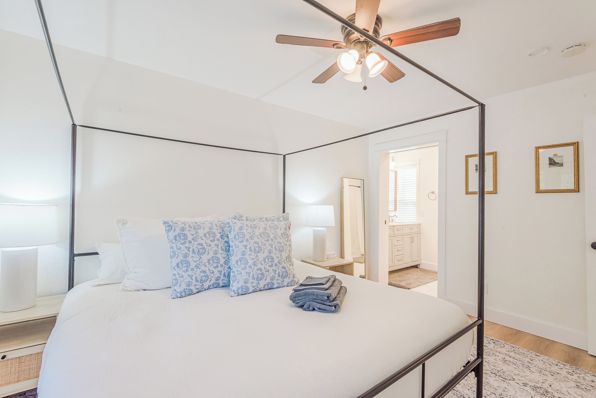 A modern bedroom with a four-poster bed, a ceiling fan, and a view into the bathroom.