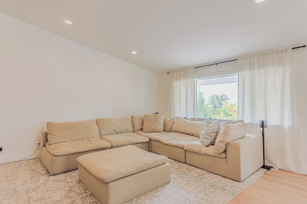 A modern living room with a beige sectional sofa, a matching ottoman, and a large window with curtains.