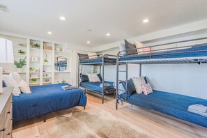 A dormitory room with bunk beds, a desk, shelves with books and plants, and a rug on the floor.