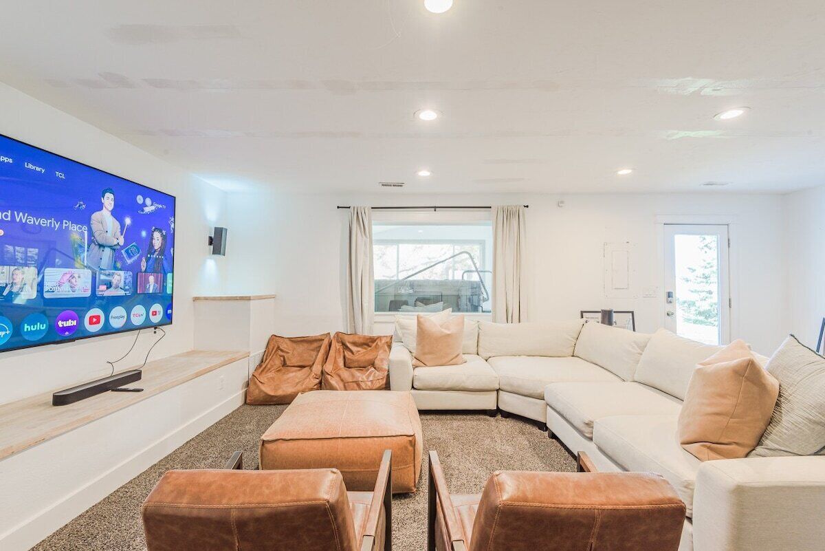 A modern living room with a large flat-screen TV, a white sofa, leather ottomans, and a window with curtains.