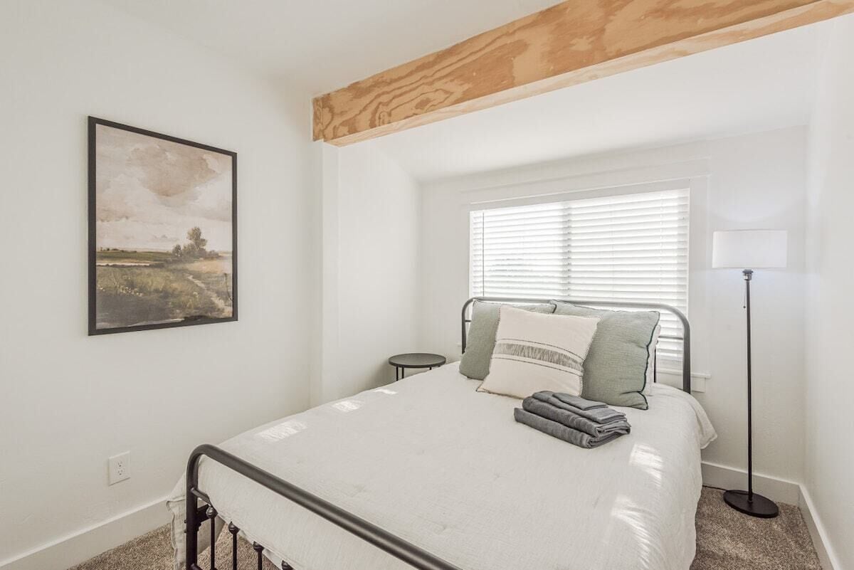 A bedroom with a bed, a bedside table, a floor lamp, a window with blinds, and a painting on the wall.