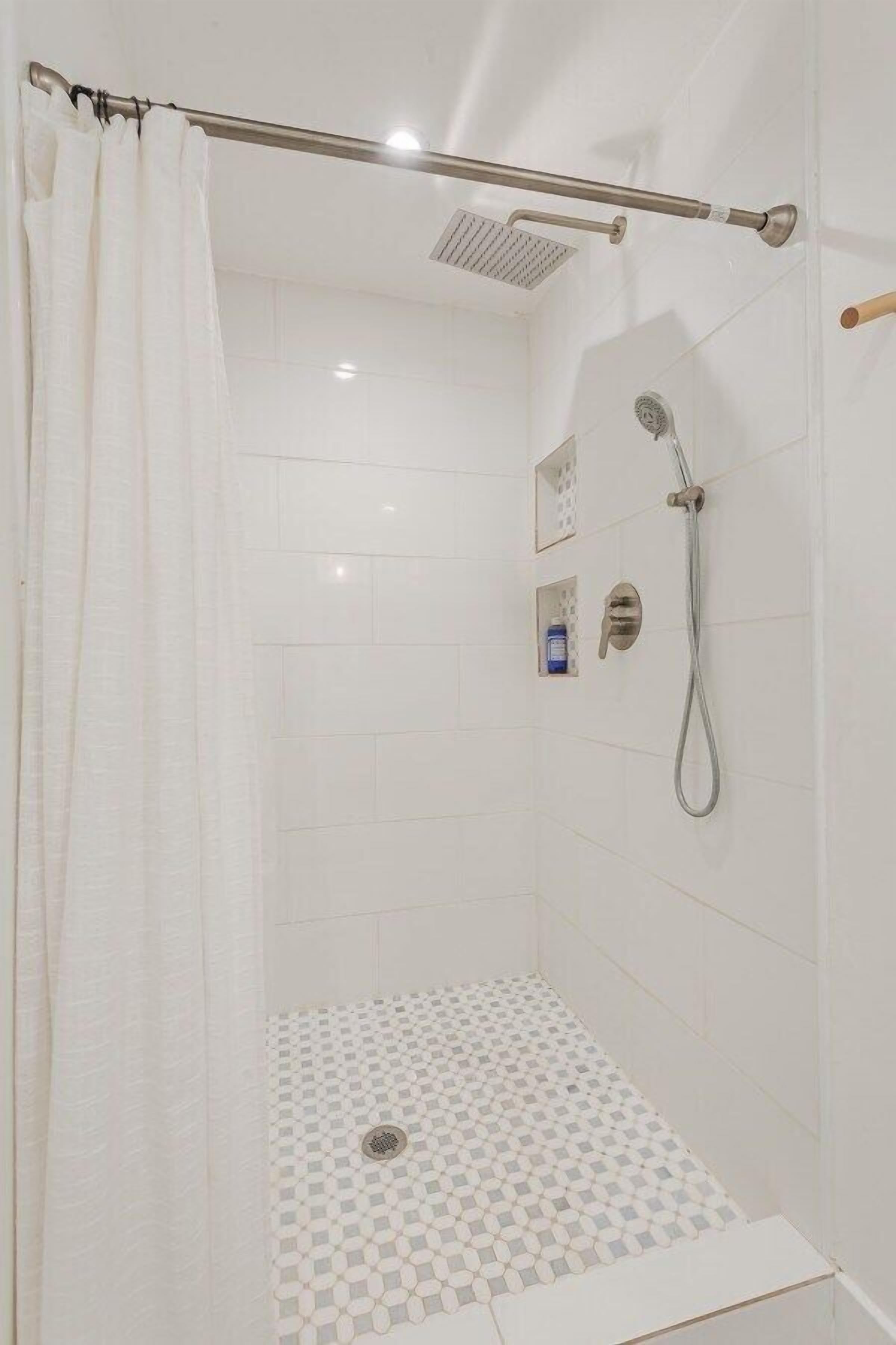 A shower area with a tiled floor, a showerhead, a control panel, and a curtain.