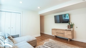 A modern living room with a flat-screen TV mounted on the wall, a wooden cabinet, and a large window with blinds.