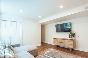A modern living room with a flat-screen TV mounted on the wall, a wooden cabinet, and a large window with blinds.