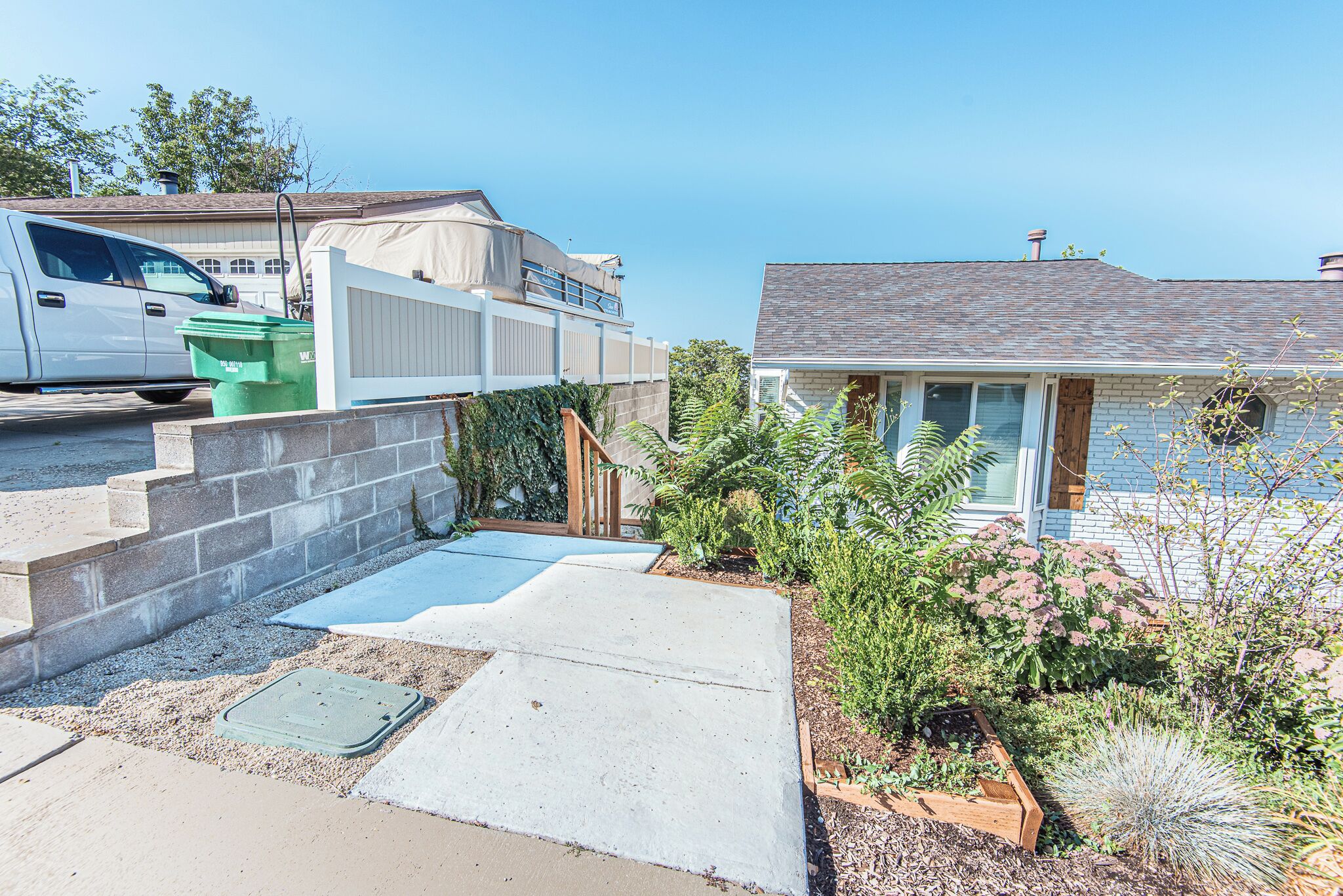 A residential driveway with a white van parked on the left, a concrete pathway leading to a house with a grey roof, and a wooden fence.