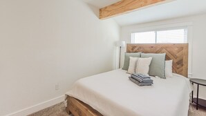 A bedroom with a wooden headboard, a window with blinds, a bedside table, and a neatly made bed with pillows and folded towels.