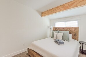 A bedroom with a wooden headboard, a window with blinds, a bedside table, and a neatly made bed with pillows and folded towels.