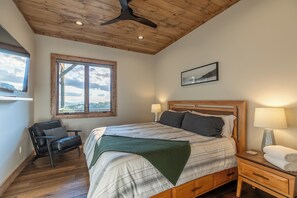 Ein Schlafzimmer mit Bett, Stuhl, Nachttisch, Lampe und einem Fenster mit Blick.