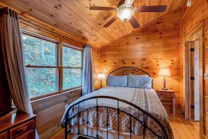Un dormitorio de cabaña de madera con una cama, un ventilador de techo y una ventana con cortinas.