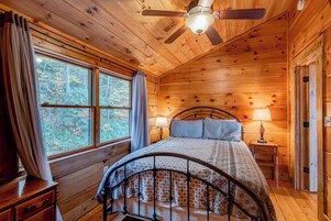 A wooden cabin bedroom with a bed, a ceiling fan, and a window with curtains.