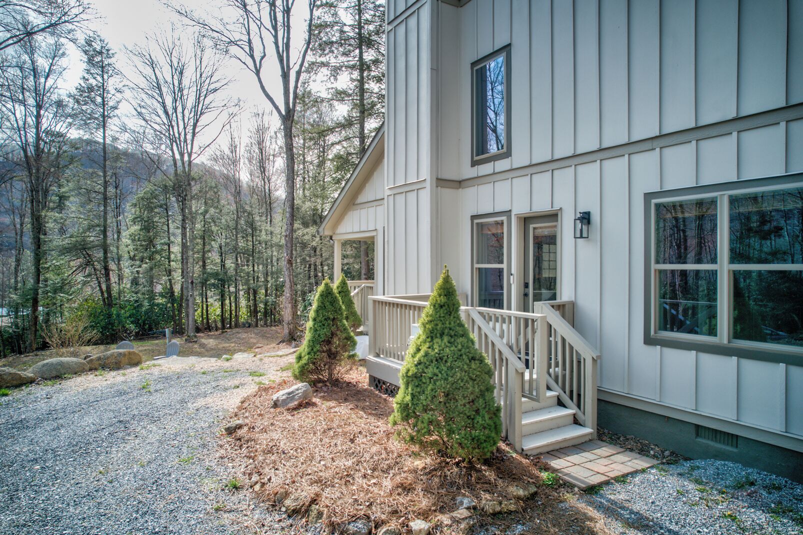 A two-story house with a covered porch, a small front yard with a gravel path, and a forested backdrop.