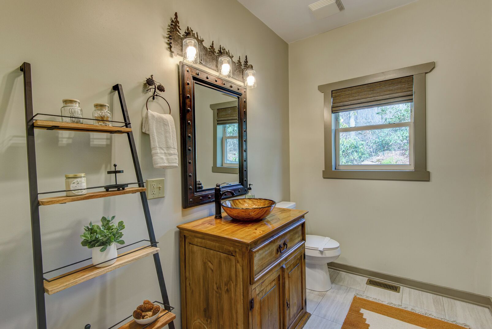 A bathroom with a wooden vanity, a large mirror with lights, a window, and shelves with jars and a plant.