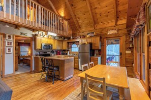A wooden cabin with a kitchen featuring a central island, bar stools, a dining table, and a view of the outdoors. - Apple Cove (Vilas)