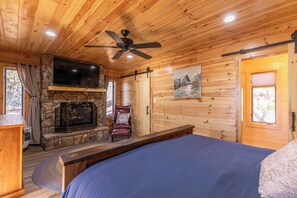 A log cabin room with a stone fireplace, a mounted TV, a bed, and a ceiling fan. - Land Harbor Lodge (Newland)