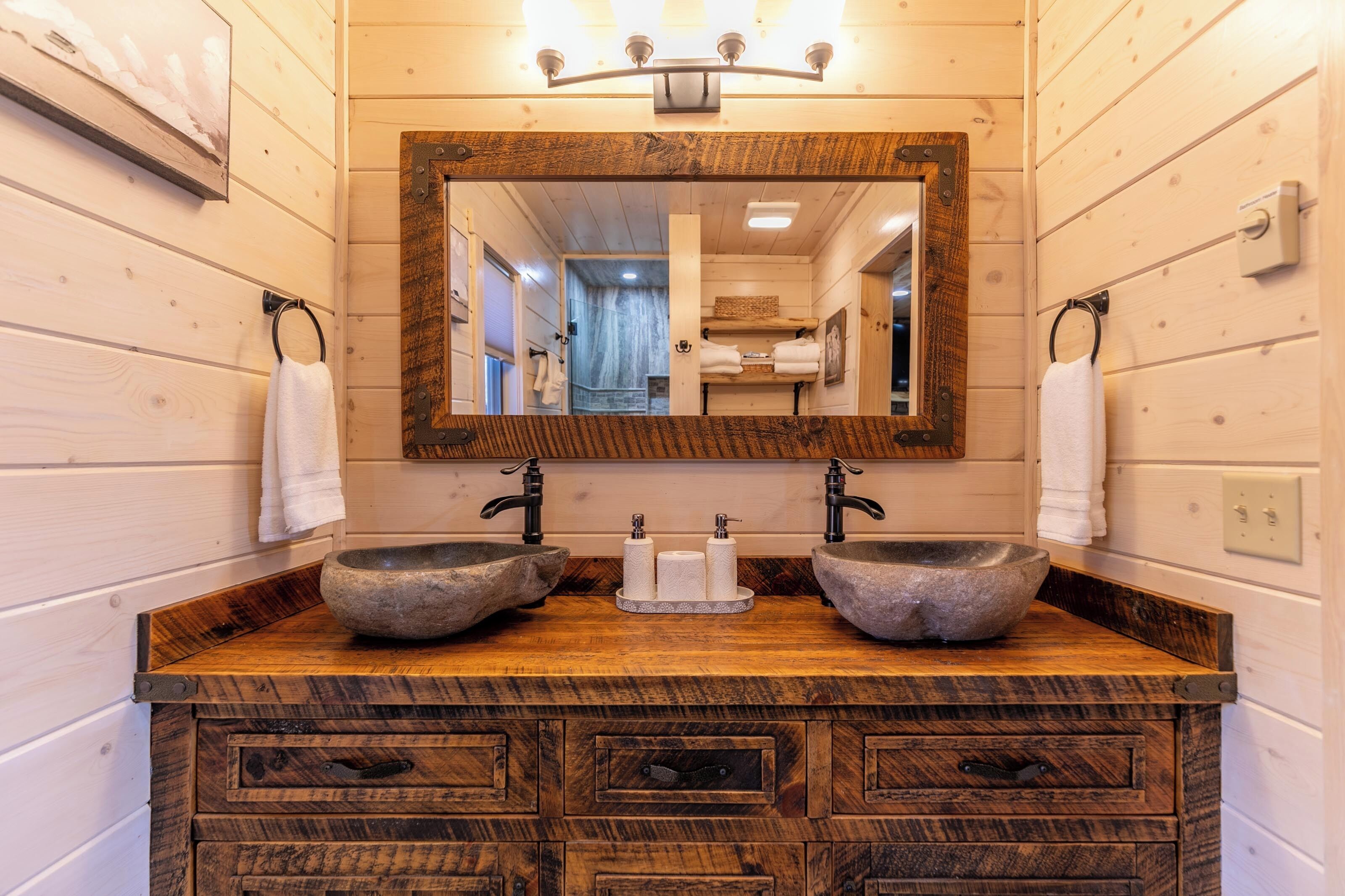 Un baño con tocador de madera, dos lavamanos de piedra, un espejo con marco de madera y argollas para toallas.