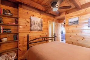 Interior de una cabaña de madera con una cama, estanterías, un cuadro y un ventilador de techo.