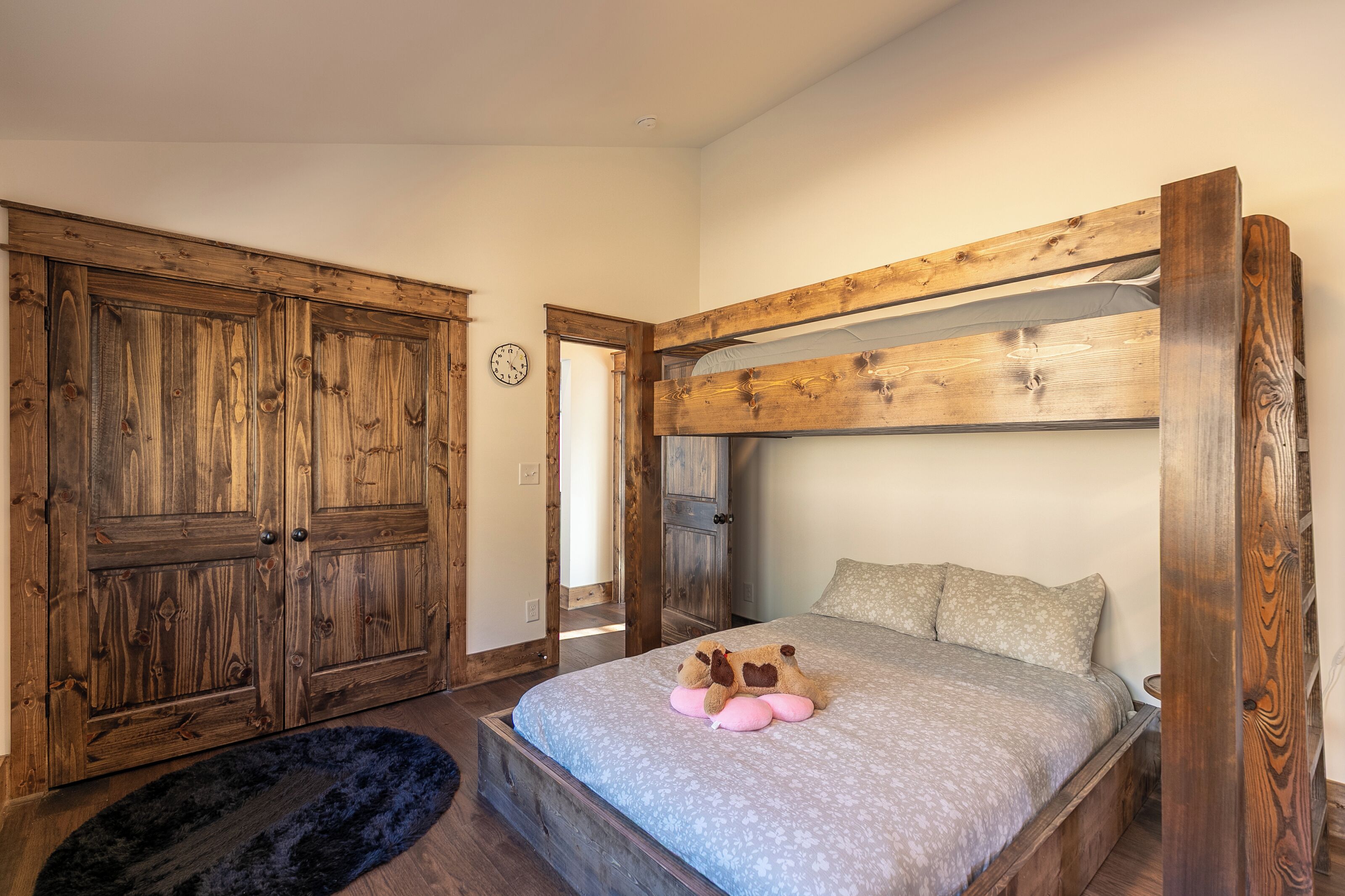 A bedroom with a wooden bunk bed, a plush bed with a patterned bedspread, a teddy bear, and a wooden wardrobe.